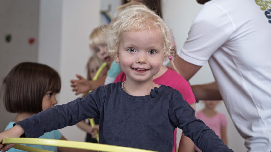Kleinkind turnt fröhlich mit dem Reifen, Trainer im Hintergrund